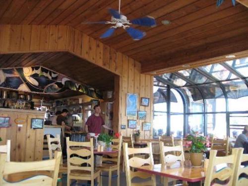 Cozy restaurant interior with wooden walls, a ceiling fan, and large windows, featuring tables with potted plants.