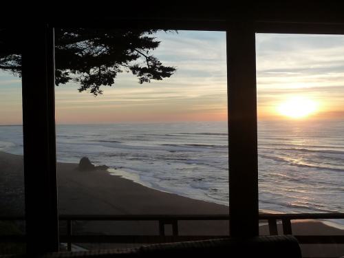 Sunset over the ocean viewed through large windows, with gentle waves and a sandy beach in the foreground.