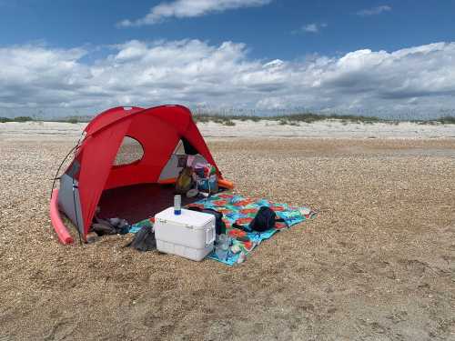 A red beach tent on a sandy shore, with a colorful blanket and cooler nearby under a partly cloudy sky.