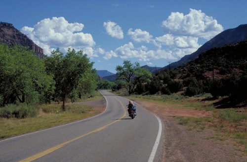 A motorcyclist rides along a winding road surrounded by green trees and mountains under a blue sky with fluffy clouds.