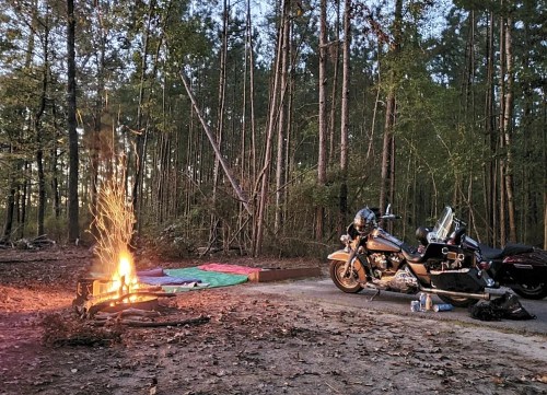 A motorcycle parked beside a campfire in a wooded area, with sleeping bags laid out on the ground.