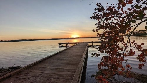 A wooden dock extends into a calm lake at sunset, with vibrant autumn leaves in the foreground.