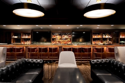 A stylish bar with a dark wood counter, shelves of liquor, and plush seating in a dimly lit lounge.