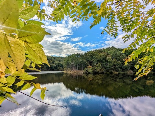 A serene lake surrounded by trees, reflecting a blue sky with clouds, framed by green and yellow leaves.