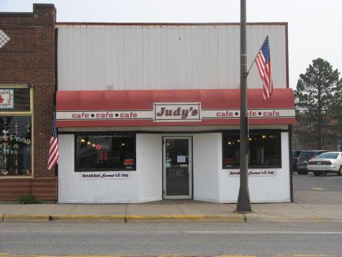 A small café named "Judy's" with a red awning, flags, and a sign stating "Breakfast Served All Day."