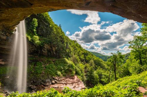 A waterfall cascades from a rocky overhang into a lush green valley under a bright blue sky with fluffy clouds.