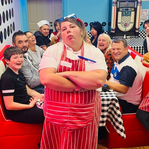 A waitress in a striped apron stands confidently in front of a lively diner group enjoying their meal.