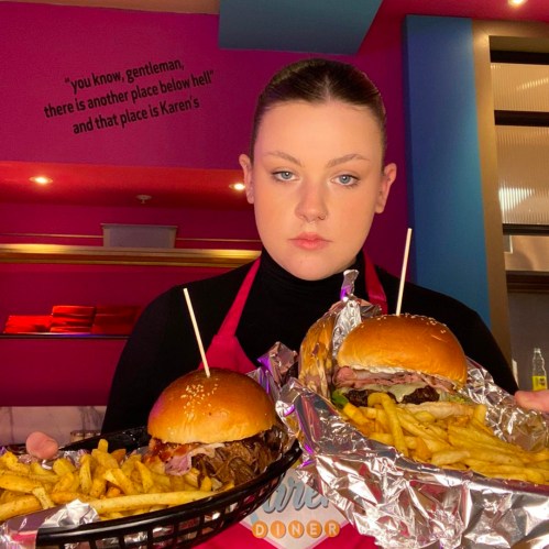 A server in a diner holds two burgers and fries, with a colorful background and a quote on the wall behind her.