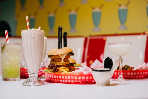 A vibrant diner table featuring a burger with toppings, milkshake, cocktails, and colorful decor in the background.