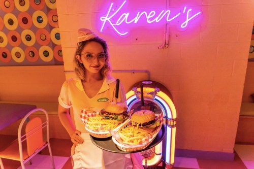 A waitress in a retro outfit holds a tray with burgers and fries, standing near a neon sign that says "Karen's."