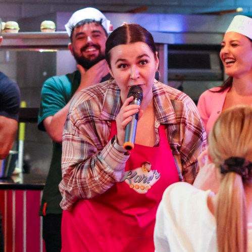 A woman in a red apron speaks into a microphone, while others smile and watch in a lively kitchen setting.