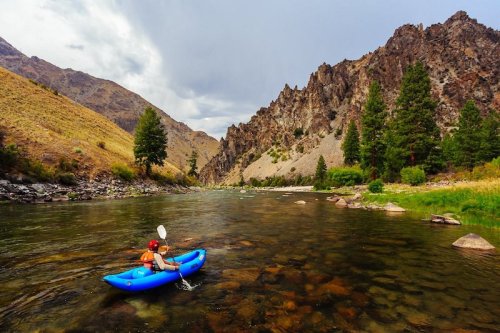 A person in a blue kayak paddles on a calm river surrounded by rocky mountains and lush greenery.