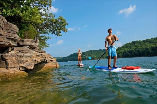 Two people paddleboarding on a calm lake, surrounded by rocky cliffs and lush greenery under a clear blue sky.