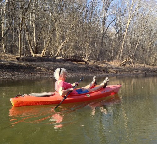 A person in a pink shirt relaxes in a red kayak on a calm river, surrounded by bare trees.