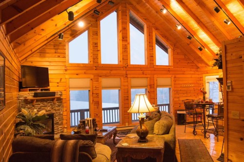 Cozy wooden cabin interior with high ceilings, a stone fireplace, and a warm ambiance from lamps and natural light.