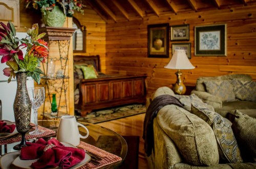 Cozy wooden cabin interior with a sofa, bed, decorative flowers, and a dining table set for two.