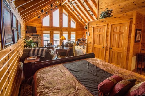 Cozy log cabin interior featuring a bedroom, living area, and large windows with a scenic view. Warm wood accents throughout.