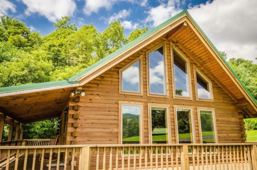 A wooden cabin with large windows, surrounded by lush greenery and a blue sky with clouds.