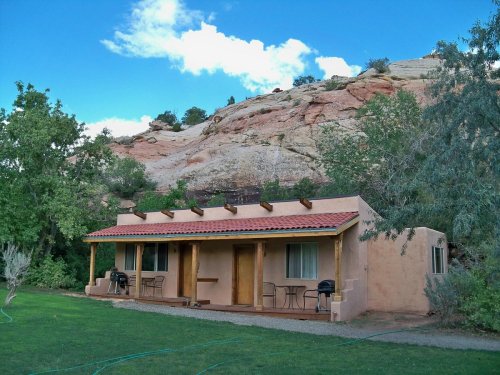 A small adobe-style house with a red tile roof, set against a rocky hillside and surrounded by green grass and trees.