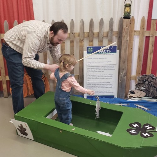 A man helps a young girl fish from a small green boat at a fun, educational exhibit.
