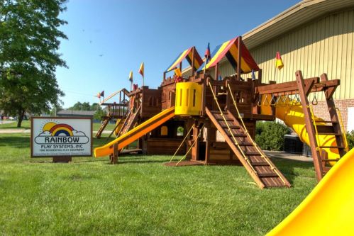 Colorful wooden playground structure with slides and flags, next to a sign for Rainbow Play Systems, Inc.