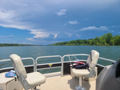 View from a boat on a lake, featuring two empty seats and a scenic backdrop of trees and clouds.