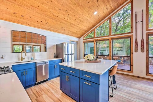 Bright kitchen with blue cabinets, white countertops, and large windows overlooking a wooded area. Modern and inviting design.