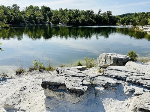 A serene lake surrounded by greenery, with rocky shores and calm water reflecting the trees.
