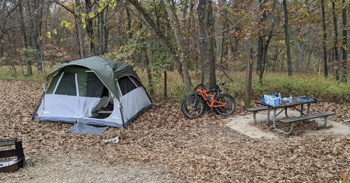 A camping scene with a tent, a mountain bike, and a picnic table surrounded by autumn leaves and trees.