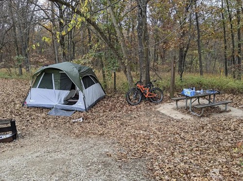 A camping scene with a tent, a mountain bike, and a picnic table surrounded by autumn leaves and trees.