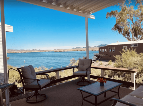 A serene lakeside view from a porch with two chairs and a table, surrounded by greenery and mountains in the distance.