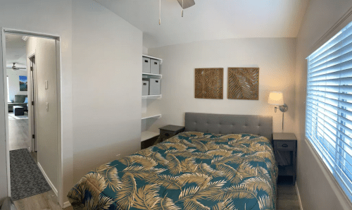 Cozy bedroom with a queen bed, palm leaf bedding, nightstands, and wall art, featuring a bright window and shelves.
