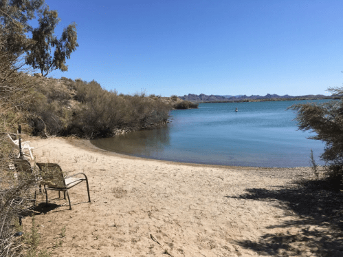 A serene beach scene with sandy shore, calm water, and distant mountains under a clear blue sky.