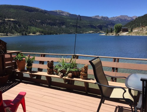 A scenic lakeside view from a wooden deck, featuring potted plants and carved bear decorations under a clear blue sky.