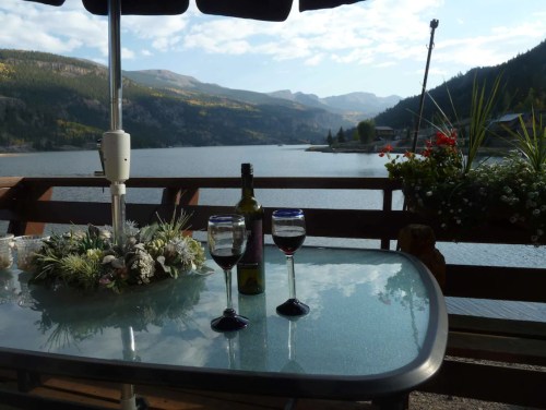 A scenic view of a lake with mountains in the background, featuring a table with two glasses of wine and a bottle.