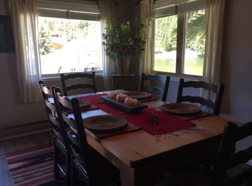 A cozy dining room with a wooden table set for a meal, surrounded by chairs and large windows overlooking a scenic view.