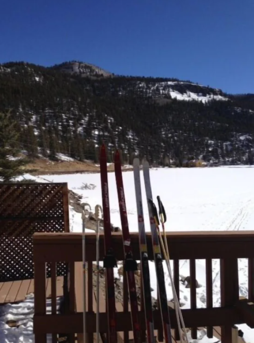 Ski poles stand on a wooden deck with a snowy landscape and mountains in the background under a clear blue sky.