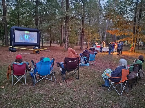 A group of people sitting in camping chairs watching a movie on an inflatable screen in a wooded area.