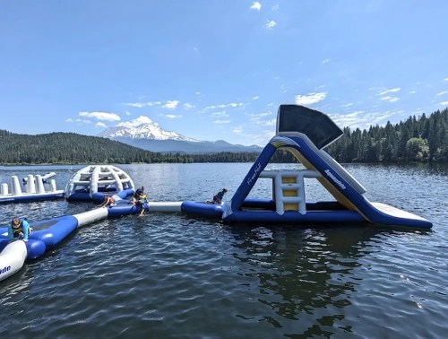 Inflatable water park on a lake with mountains in the background; people enjoying the attractions under a clear blue sky.