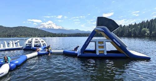 Inflatable water park on a lake with mountains in the background; people enjoying the attractions under a clear blue sky.