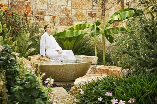 A woman in a white robe sits by a stone basin in a lush garden with tropical plants and colorful flowers.