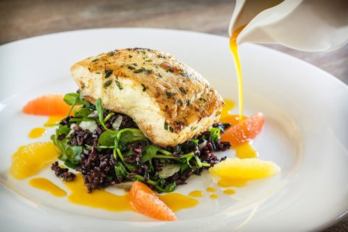 A beautifully plated dish featuring a piece of fish on a bed of greens and black rice, drizzled with a citrus sauce.