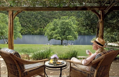 A woman sits on a porch by a lake, enjoying tea and breakfast with a scenic view of greenery and water.