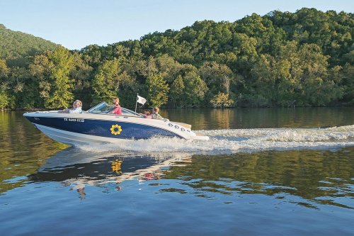 A boat with four people speeds across a calm lake, surrounded by lush green trees and reflecting water.