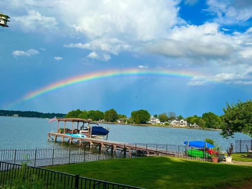 A vibrant rainbow arcs over a calm lake, with a dock and colorful boats in the foreground and lush greenery nearby.