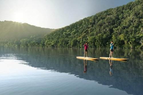 Two people paddleboarding on a calm lake surrounded by lush green hills under a bright sky.