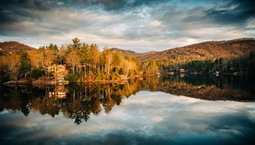 A serene lake reflects trees and a house, surrounded by mountains under a cloudy sky at sunset.