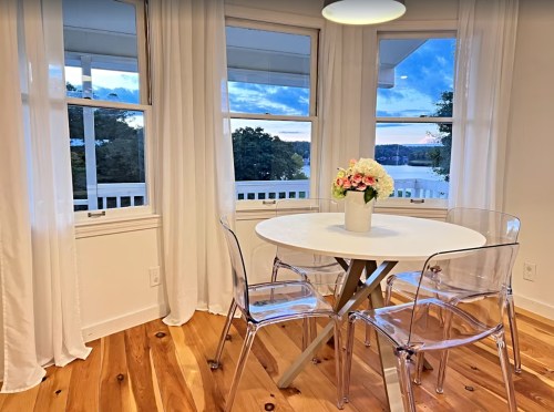 Bright dining area with a round table, clear chairs, and a vase of flowers, overlooking a scenic view through large windows.