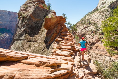 A hiker in a blue shirt and pink shorts walks along a rocky trail in a canyon surrounded by steep cliffs and greenery.