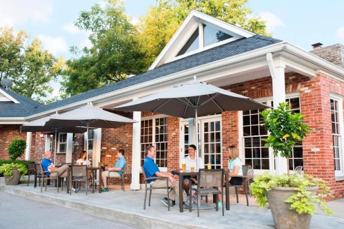 A group of people enjoying drinks at outdoor tables under umbrellas in a sunny patio setting.